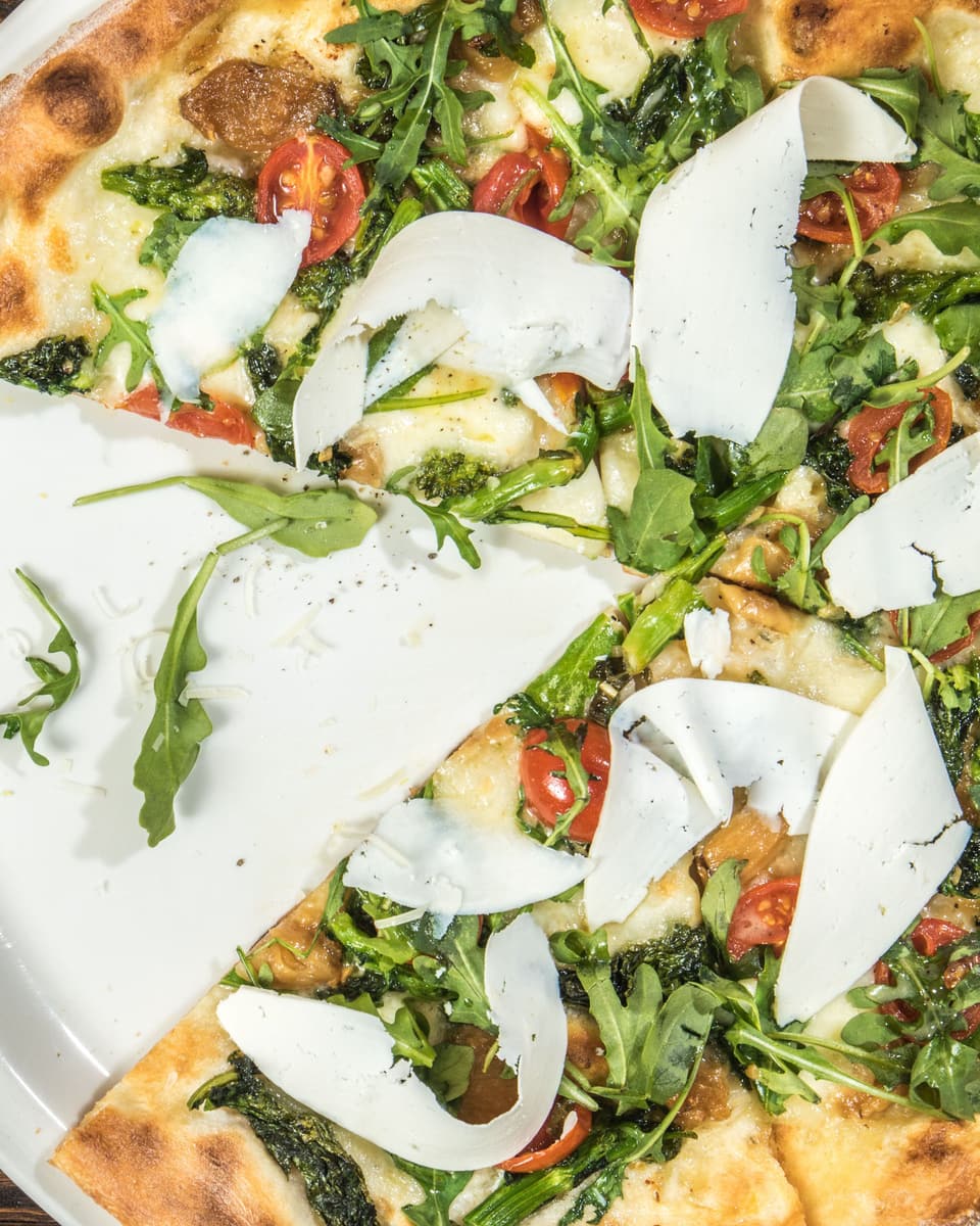 A wood-fired flatbread with greens, tomatoes and shaved cheese, two slices already gone