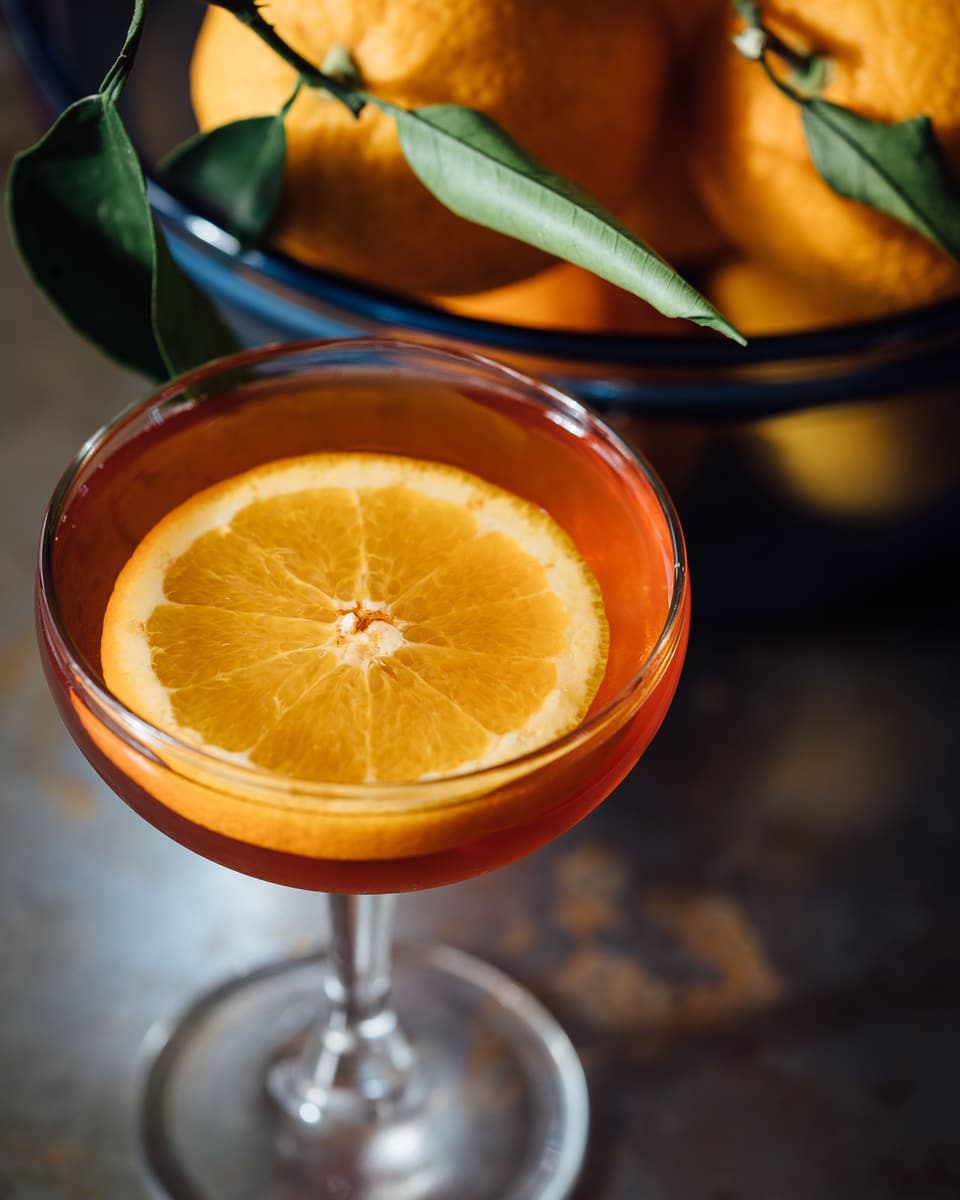 An orange drink in a coupe glass with a slice of orange on it, a bowl of oranges behind