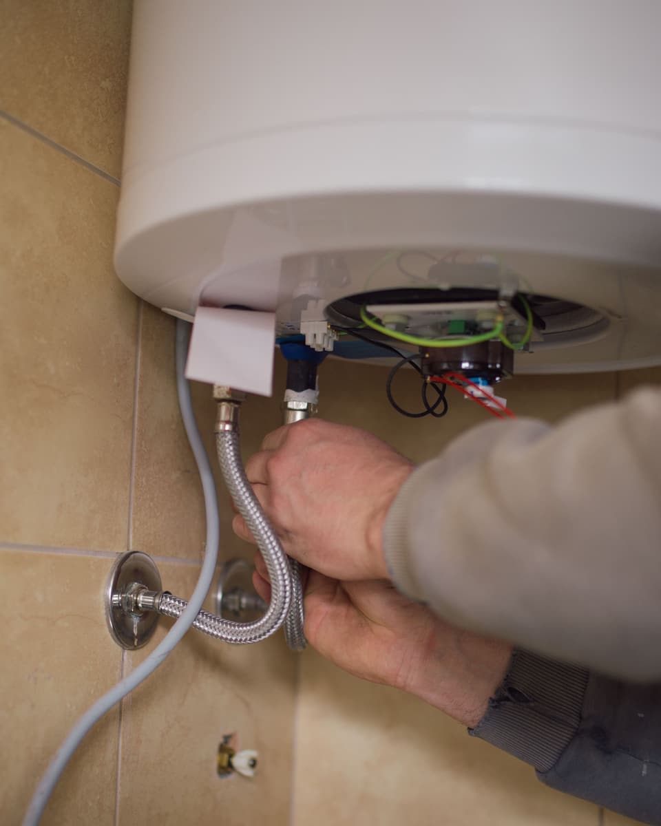 Two hands fitting a braided supply hose under a wall-hung water heater.
